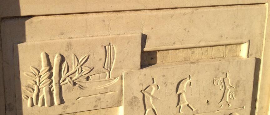Tall apartment buildings going up next to vacant lots. In the foreground, ancient scenes carved into a stone railing.