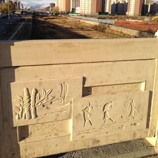 Tall apartment buildings going up next to vacant lots. In the foreground, ancient scenes carved into a stone railing.