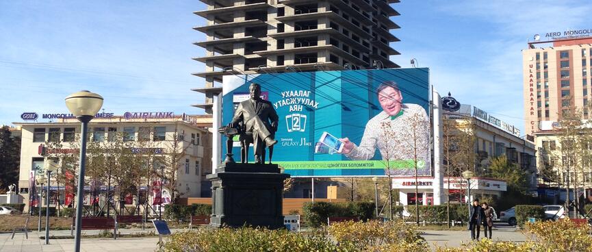 A man on a billboard seems to reach out towards a metal statue of a man in a chair.