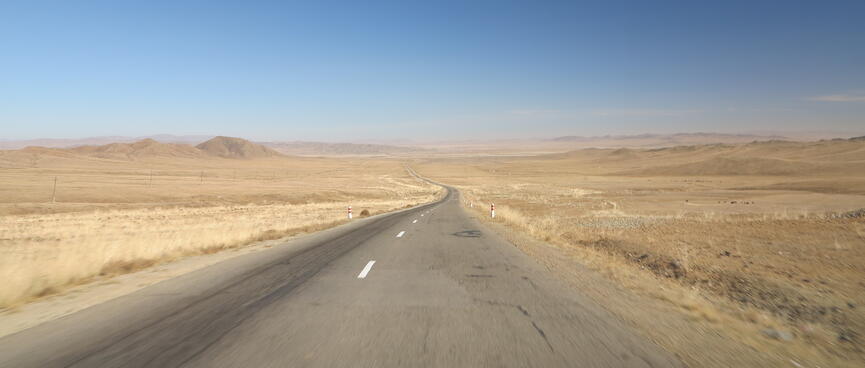 A grey two lane highway stretches through a light brown landscape.