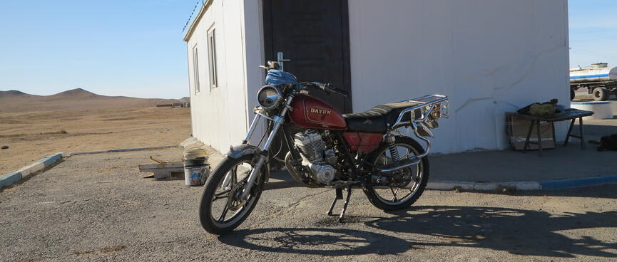 A red Dayun motorcycle parked outside a square white building with blue trim.
