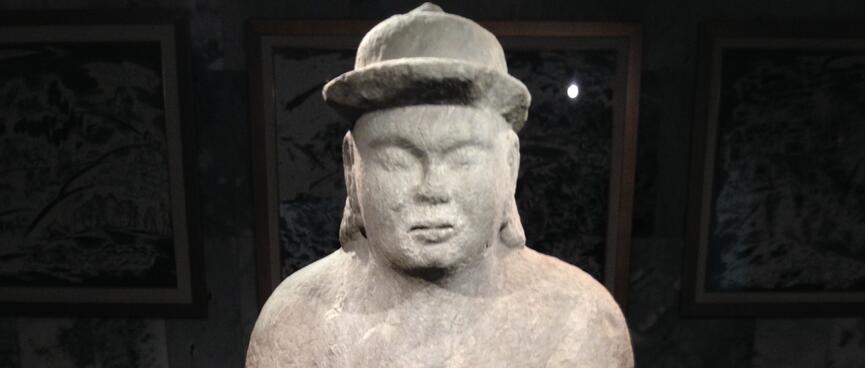 A stone man wears a bowler hat and holds a cup in one hand.