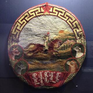 A felt badge showing a rider on a galloping brown horse, under a bright sun. Four other animals are featured on the border of the badge.
