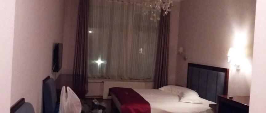 A pinky hued room containing a white bed with a red runner, and a fancy chandelier.