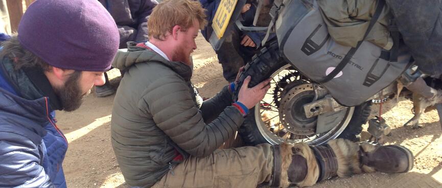 One of the motorcycle tourists fits his back wheel while the other looks on.