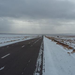 A two lane asphalt highway is surrounded by snow-covered ground and grey skies.
