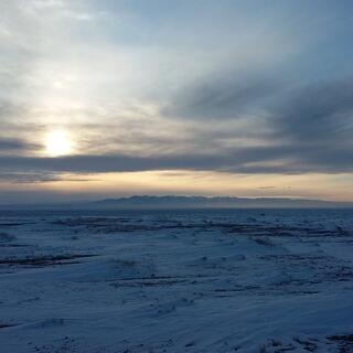 A pale yellow sun lights up a mountain in the distance, while in the foreground grey clouds colour the steppe dark blue.