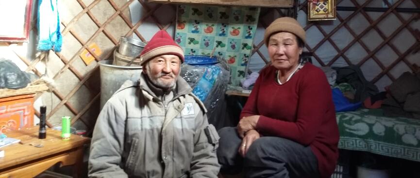 A man with a grey beard and a red hat sits next to a woman with a brown hat. Both are smiling ever so slightly.