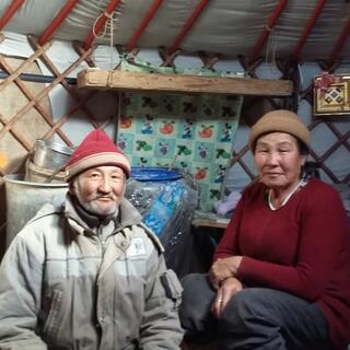 A man with a grey beard and a red hat sits next to a woman with a brown hat. Both are smiling ever so slightly.