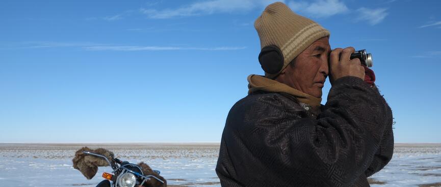 A man with a black jacket, brown hat and ear muffs surveys the landscape with my monocular. His motorcycle is parked behind him.