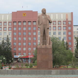 A large statue of Lenin, on a pedestal outside an eight story building with many windows, in Chita.