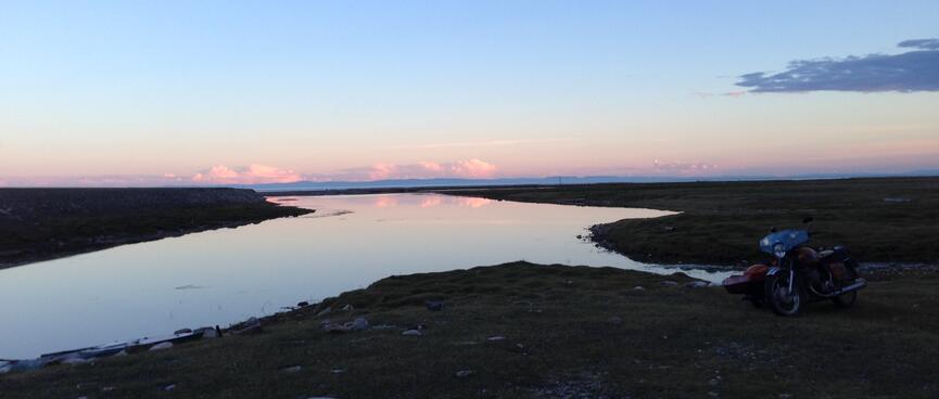 A motorcycle stands next to an inlet at sunset.