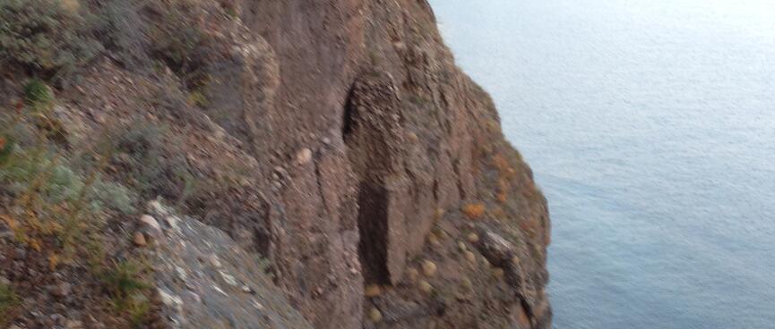 The trail clings to the cliffside, above the opening to a cave just above the waterline.