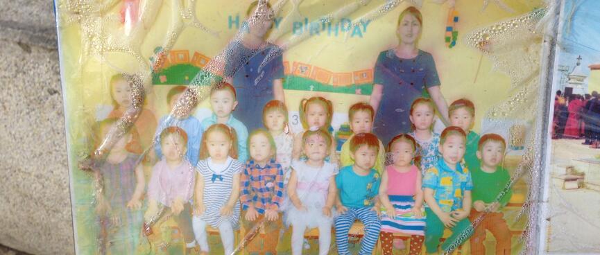 Class photo shows a group of small children and two teachers in blue dresses.
