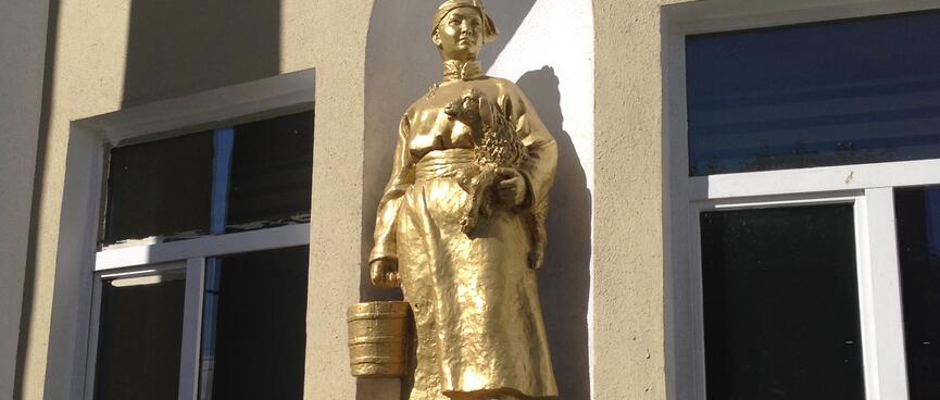 A golden statue of a woman in traditional Mongolian dress, carrying a lamb and a bucket.