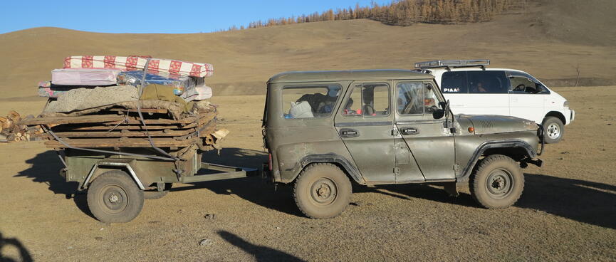 A green army jeep tows a small trailer stacked with belongings.
