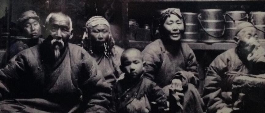 A black and white photo of a Mongolian family seated on the floor with cooking utensils behind them.