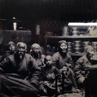 A black and white photo of a Mongolian family seated on the floor with cooking utensils behind them.