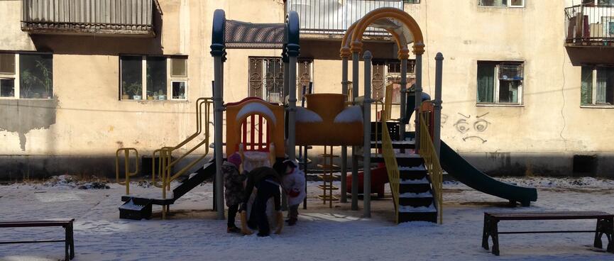 A parent clears snow off a playground slide while two children wait.