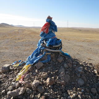 A wooden pole on top of a pile of rocks. It is wrapped in coloured scarves and plastic or rubber tubes.