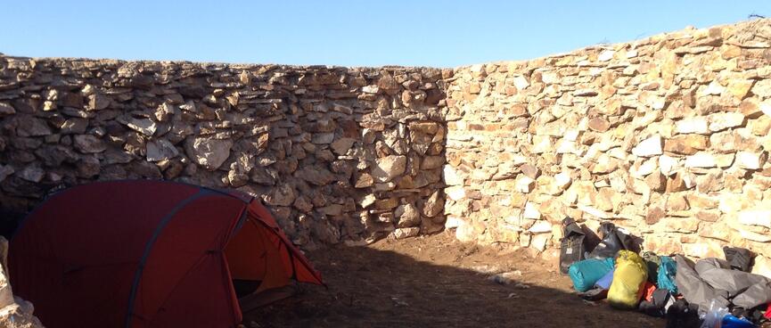 My orange tent and a stack of bags in the corner of a stone-walled compound with a dirt floor.