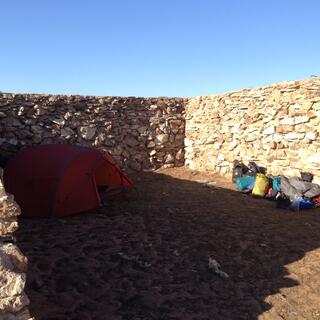 My orange tent and a stack of bags in the corner of a stone-walled compound with a dirt floor.