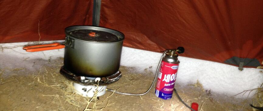 A camping pot sits on a gas cooker in the corner of my tent. The gap between the tent and the ground is packed with snow.