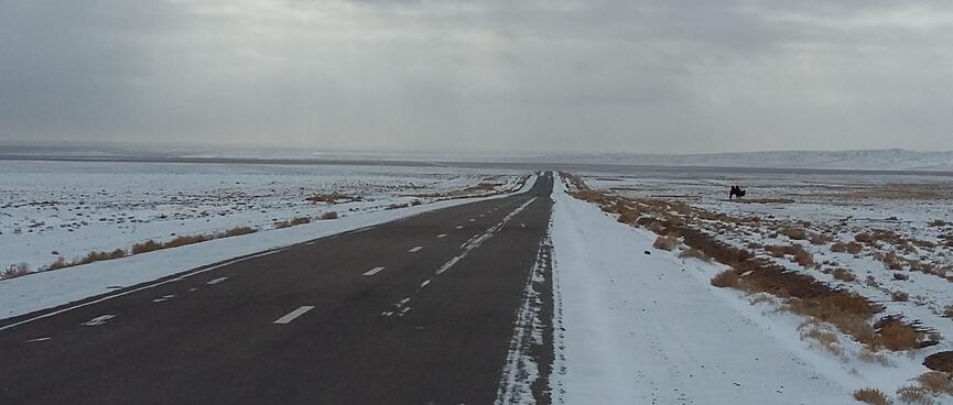 A two lane asphalt highway is surrounded by snow-covered ground and grey skies.