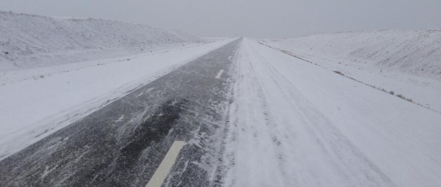 Snow blows over the mostly snowless lane of a two lane highway.