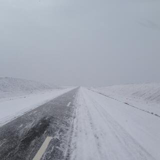 Snow blows over the mostly snowless lane of a two lane highway.
