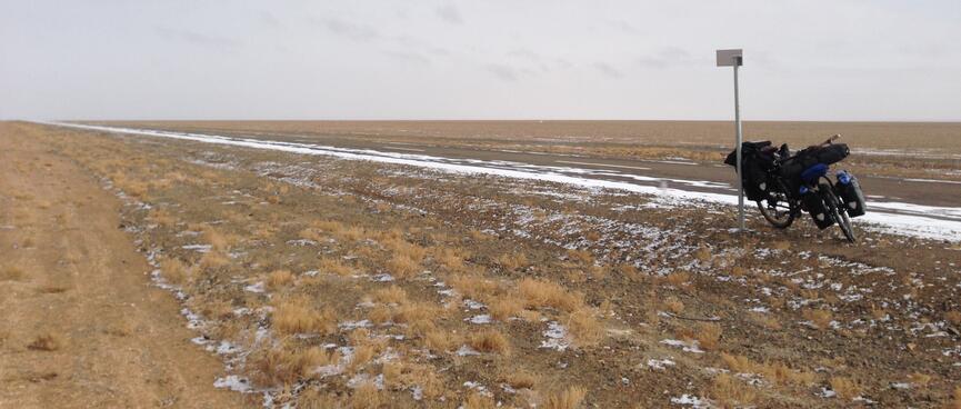 My loaded bike rests against a signpost. The browny golden steppe is mostly clear except for a little ice.