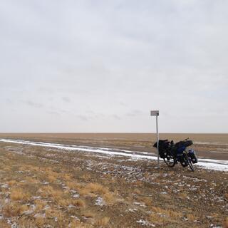 My loaded bike rests against a signpost. The browny golden steppe is mostly clear except for a little ice.