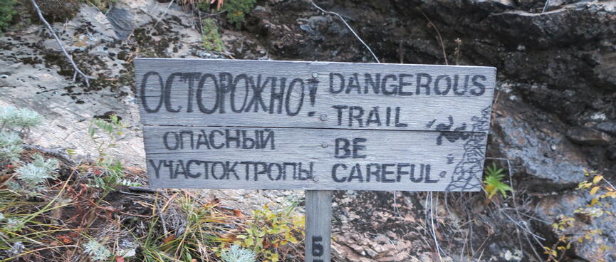 A bilingual wooden sign shows a hiker falling down a rocky bank.